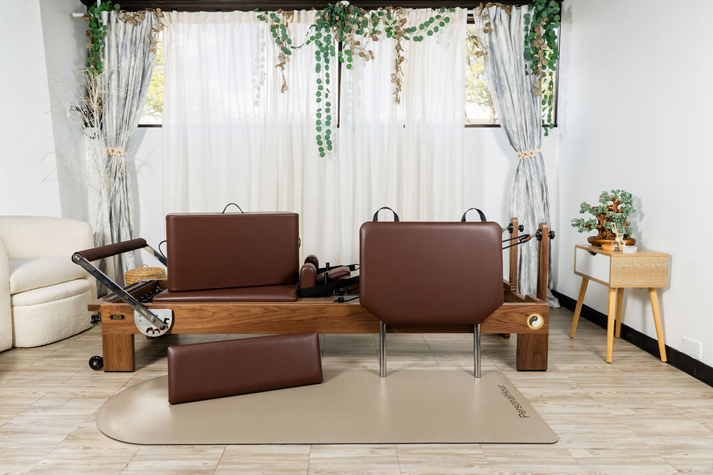 Brown Pilates reformer with cushions, a chair, and an accent table on a wooden floor with white curtains and plants in the background.